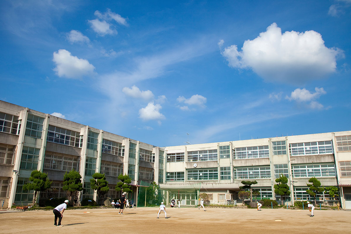 緑に囲まれた中学校の広いグラウンド。奥には校舎と山が見える。